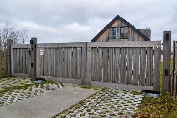 Een vergrijsde houten poort van Padouk met een woning op de achtergrond