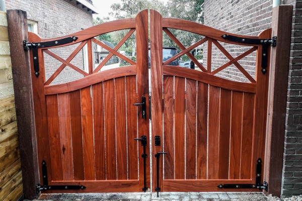 Dubbele padouk tuinpoort met gebogen bovenzijde, kruisdetails en zwart hang- en sluitwerk.